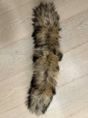 Pälskrage äkta raccoon tvättbjörn - Säljer en fluffig sjal i äkta päls med beige, bruna och svarta toner. Sjalen har en långsmal form och ger en lyxig känsla till din outfit. Perfekt att slänga över axlarna för extra värme och stil eller fästa på din luva. 70 cm. Köpt på Vivaldi. 