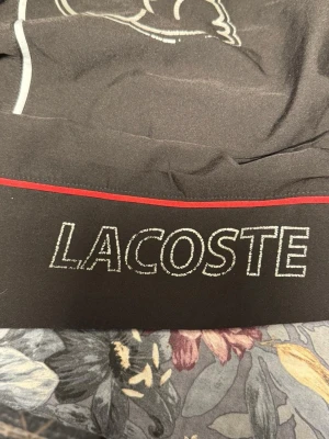 Svart vindjacka från Lacoste - Snygg svart vindjacka från Lacoste med stor krokodil-logga och röd detalj på ryggen. Jackan har huva, resår i midjan och ärmar samt vita och röda linjer längs ärmarna. Perfekt för dig som vill sticka ut med en sportig och trendig look.