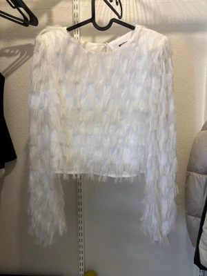 Vit fluffig topp med långa ärmar - Säljer en superfin vit topp med långa ärmar och fluffig, fjäderliknande textur. Toppen är croppad och har en unik struktur som verkligen sticker ut. Tröjan har vadderade axlar som får axlarna att poppa. Perfekt för dig som vill ha något annorlunda och trendigt i garderoben.