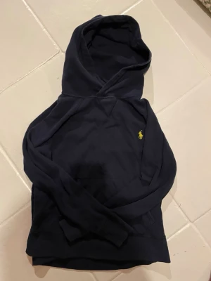 Mörkblå hoodie från Ralph Lauren - Mörkblå hoodie från Ralph Lauren med klassisk gul broderad logga på bröstet. Hoodien har en stor huva och magficka framtill. Tillverkad i mjukt bomullsmaterial som är skönt att bära. Perfekt för en avslappnad och stilren look. i grymt skick och är storlek XS kom om du har minsta lilla fråga! 📦🔥