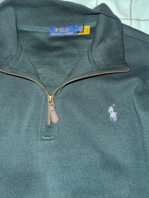 Mörkgrön half-zip från Polo Ralph Lauren - Mörkgrön långärmad tröja från Polo Ralph Lauren med klassisk half-zip och dragkedja i metall med läderdetalj. Broderad lila logga på bröstet. Tröjan har ribbade muddar och är tillverkad i mjukt bomullsmaterial.