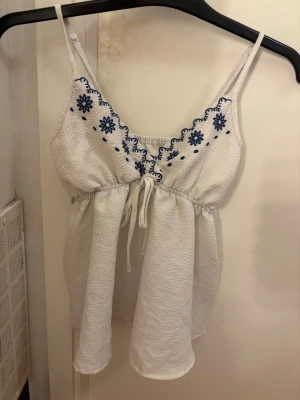 Vit topp med blå broderi och knytband - Söt vit topp med tunna axelband och blå broderade blommor längs halsringningen. Toppen har ett knytband under bysten och är tillverkad i ett lätt, strukturerat tyg som ger en luftig känsla. Perfekt för sommarens varma dagar.