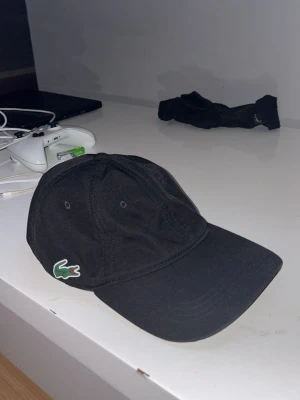 Svart keps från Lacoste - Snygg svart keps från Lacoste med klassisk krokodillogga broderad på sidan. Kepsen har böjd skärm och justerbar rem baktill. Perfekt accessoar för en sportig och stilren look.