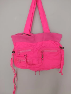 Knallrosa handväska med dragkedjor - Cool knallrosa handväska i nylon med flera dragkedjefickor framtill och axelband. Väskan har detaljer som spännen och dekorativa remmar samt ribbad kant upptill. Den går att förstora genom en dragkedja som sitter på utsidan av väskan. Har flera fack på insidan. Perfekt för dig som gillar färgstarka accessoarer och vill sticka ut. Väskan är i fint använt skick, har några små märken på utsidan som knappt syns på bild, samt ett märke på insidan. Märket Kookai