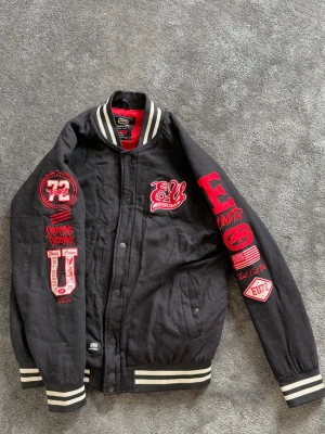 Svart bomberjacka från Ecko Unlimited - Snygg svart bomberjacka från Ecko Unlimited med broderade patches och tryck i rött och vitt på både ärmar, bröst och rygg. Jackan har ribbade muddar och krage med vita ränder samt två fickor fram. Perfekt för dig som gillar streetwear och vill sticka ut.