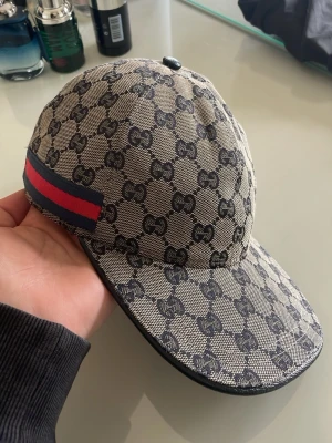 Gucci beige keps med GG-mönster - Säljer en beige keps från Gucci med klassiskt GG-monogram i svart över hela kepsen. På sidan finns en ikonisk röd och marinblå rand. Kepsen har böjd skärm och är tillverkad i canvasmaterial som ger en exklusiv känsla.