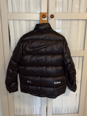 Svart pufferjacka från Nike NOCTA - Snygg svart pufferjacka från Nike NOCTA med glansig finish och broderade loggor på bröstet. Jackan har hög krage, dragkedja framtill och två sidofickor. Perfekt för kalla dagar när du vill hålla stilen. Kan gå ner i pris vid snabb affär