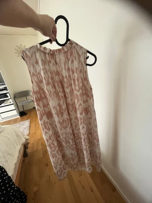 Ljusmönstrad ärmlös klänning - Säljer en luftig och ärmlös klänning med ljusrosa och vita abstrakta mönster. Klänningen har en rak passform och rundad halsringning, perfekt för varma dagar. Materialet känns lätt och svalt, troligen polyester eller bomull.