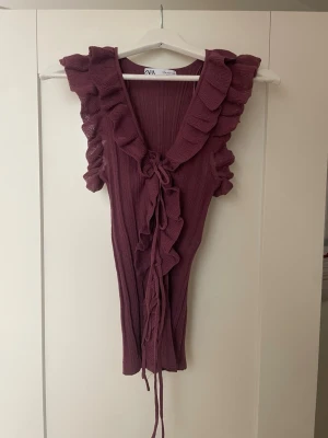 Vinröd volangtopp från Zara - Supersöt vinröd topp från Zara med volangkant längs urringningen och snörning framtill. Toppen är ärmlös och har en ribbad struktur som ger en snygg passform. Perfekt för dig som vill ha något extra till outfiten.