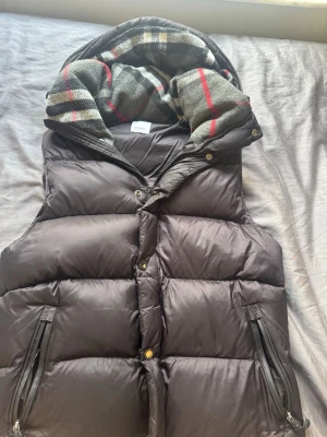 Svart dunväst från Burberry - Snygg svart dunväst från Burberry med avtagbar huva fodrad i klassiskt rutigt mönster i grått, rött och beige. Västen har dragkedja och tryckknappar framtill samt två fickor med dragkedja. Perfekt för lager på lager och streetstyle.