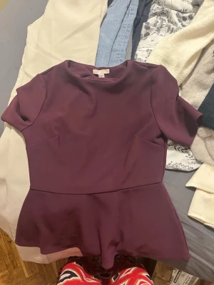 Mörklila peplum topp - Säljer en mörklila peplum topp med korta ärmar och rund halsringning. Toppen har en figurnära passform upptill och en volang nedtill som ger en snygg siluett. Perfekt att matcha med jeans eller kjol för en trendig look.