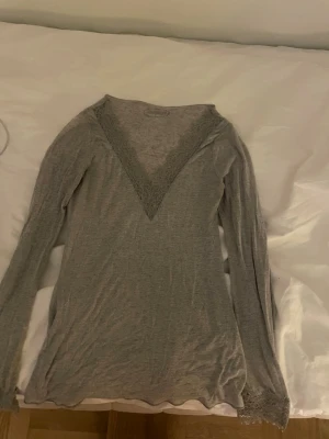 Grå topp från Intimissimi - OBS DEN HAR TVÅ HÅL PÅ RYGGEN (kanske går att rädda ifall man syr eller liknande) Säljer en grå långärmad topp från Intimissimi med djup V-ringning dekorerad med spetsdetaljer. Tillverkad i mjukt och stretchigt material som passar perfekt för avslappnade dagar hemma.