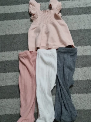 Sött stickat set med volangärmar - Stickat set med ljusrosa topp med volangärmar och tre par ribbade byxor i rosa, vitt och grått. Toppen har en lös passform och mjukt material, perfekt för en bekväm och söt look. Inga synliga märken.