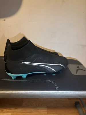 Svarta Puma fotbollsskor utan snörning - Säljer ett par svarta fotbollsskor från Puma Elite i storlek 43 använt dem en gång men de passade mig inte.  Skorna har en strumpliknande passform och vita detaljer längs sidan samt ljusblå dobbar. Perfekta för dig som vill ha bra grepp och kontroll på planen.
