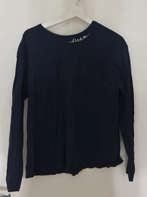 Mörkblå långärmad topp med text - En enkel mörkblå långärmad topp med rund halsringning och vit text 'dream' tryckt nära kragen. Tunn och mjuk bomullskänsla, perfekt för att matcha med jeans eller kjol. Stilren och avslappnad design.