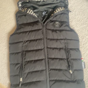 Svart dunväst från Philipp Plein - Snygg svart dunväst från Philipp Plein med quiltad design och dragkedja framtill. Västen har huva med logotypdetaljer och två fickor med dragkedja. INTE äkta men extremt realistisk. pris går att diskuter!!