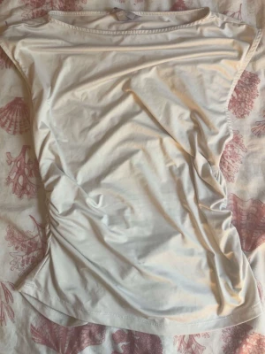 Vit ärmlös topp med rynkade sidor - Säljer en enkel och stilren vit topp med ärmlös design och rynkade sidor för en snygg passform. Perfekt till jeans eller kjol och lätt att matcha med olika stilar. Mjuk och skön mot huden.