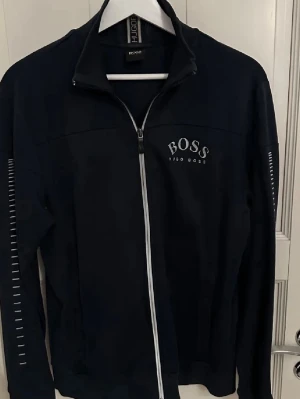 Mörkblå mjukisdress från Hugo Boss - Säljer en mörkblå mjukisdress från Hugo Boss med logga tryckt på benet och elastisk midja med Hugo Boss-tryck. Byxorna har en avslappnad passform och är tillverkade i mjukt material som passar perfekt för chill dagar. Snygg och stilren design med diskreta detaljer. Zip hoodie är i storlek S, Byxorna är i storlek M. Paketpriset landar på 1500kr