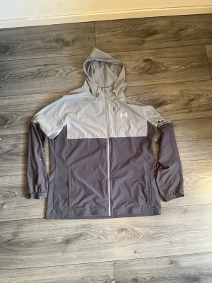  Under Armour  - Snygg tvåfärgad grå vindjacka från Under Armour med huva och hel dragkedja framtill. Jackan har logga på bröstet, elastiska muddar och praktiska sidofickor. Perfekt för blåsiga dagar och sportiga outfits.