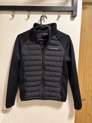 Svart cardigan från Peak Performance - Snygg svart jacka från Peak Performance med quiltad front och rygg samt släta ärmar. Jackan har dragkedja framtill, hög krage och två sidofickor med dragkedja. Perfekt för lager-på-lager och stilren look.  Nypris 2500kr