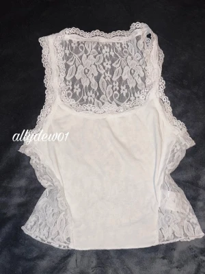 Vit spetslinnetopp med blommönster - Säljer en vit ärmlös linnetopp med transparent spets och blommigt mönster. Toppen har fina spetsdetaljer runt kanterna och är perfekt för lager på lager eller som statement-plagg. Materialet är mjukt och stretchigt med tydlig spetskänsla.