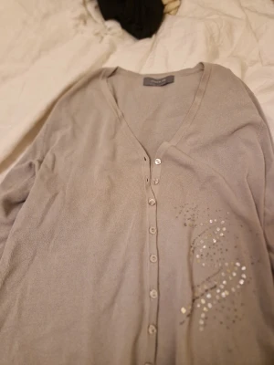 Grå cardigan från Claire.dk - Mjuk grå cardigan från Claire.dk med v-ringning och knappar framtill. Tröjan har dekorativa paljettdetaljer på ena sidan och är tillverkad i ett tunt, bekvämt material. Perfekt att slänga över en topp för en chill look.