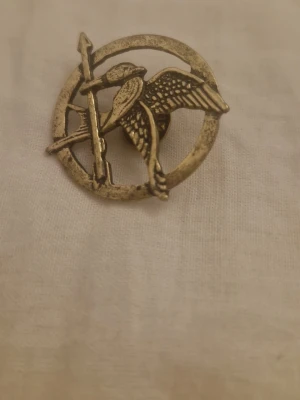 Mockingjay brosch i antikguld - Säljer en snygg brosch i antikguld med motiv av en fågel med pil och cirkel, inspirerad av Hungerspelen. Broschen är gjord i metall och har en säkerhetsnål på baksidan. Perfekt för dig som vill ha en unik accessoar med coolt motiv.