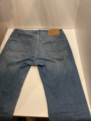 Blå Jack & Jones  med raka ben - Klassiska blå Jack & Jones  jeans med raka ben och femficksmodell. Jeansen har en ljus tvätt och läderpatch bak i midjan. Tillverkade i bomull med knappgylf och tydliga sömmar på bakfickorna.