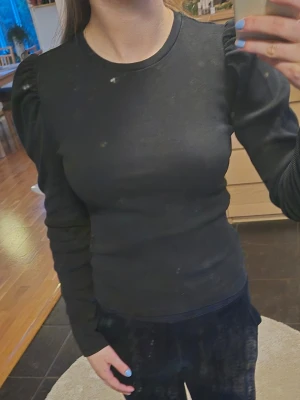 Svart långärmad topp med puffärm - Snygg svart långärmad topp med puffiga axlar och rund halsringning. Modellen är figurnära och toppen är tillverkad i ett stretchigt ribbat material som ger en bekväm passform. Perfekt för dig som vill ha en enkel men trendig look.