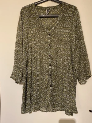 Grön mönstrad blus från Isolde - Snygg grön blus från Isolde med blommigt mönster i vitt och gult. Blusen har knappar framtill, v-ringning och trekvartsärm. Tunt och luftigt material som ger en avslappnad känsla. Perfekt för dig som gillar bohemisk stil.