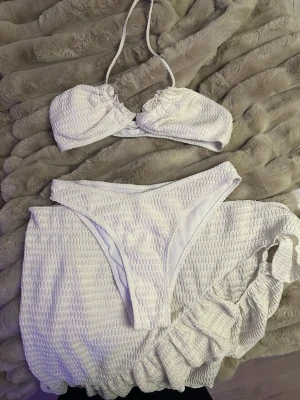 Vit bikini med struktur och kjol - Trendig vit bikini med strukturerat tyg och bandeau-topp med knyt i nacken. Matchande högt skurna bikinitrosor och en söt kjol i samma material. Perfekt för stranden eller poolen och ger en fräsch sommarkänsla.
