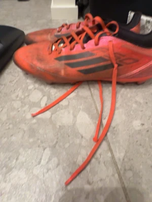 Röda Adidas fotbollsskor med dobbar - Säljer ett par röda Adidas fotbollsskor med svarta detaljer och klassiska tre ränder på sidan. Skorna har snörning och fasta dobbar för bra grepp på gräsplan. Ovandelen är i syntetmaterial och designen är sportig och modern. Perfekta för dig som vill sticka ut på planen.