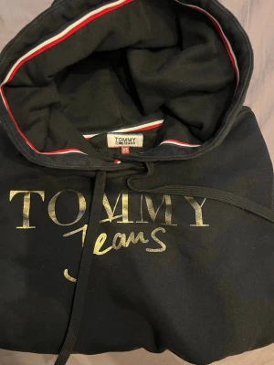 Svart hoodie från Tommy Jeans XS - Snygg svart hoodie från Tommy Jeans med guldfärgat tryck på bröstet. Klassisk huva med dragsko och rödvit detalj på insidan av huvan. Tillverkad i mjukt bomullsmaterial som är skönt att bära. Perfekt för en avslappnad och trendig look.