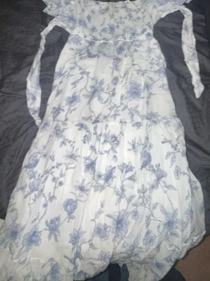 Vit långklänning med blått blommönster - Romantisk långklänning i vitt med blått blommigt mönster. Klänningen har långa ärmar och volangdetaljer, samt en luftig och flowy passform. Perfekt för sommarens alla tillfällen.