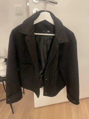 Mörkgrå kappa från 157 - Stilren mörkgrå kappa från 157 i ullmix med klassisk krage och två stora fickor framtill. Kappan är fodrad med svart tyg och har knappar vid ärmslut och framtill.