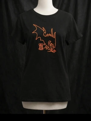 Svart t-shirt med orange drakmotiv - Cool svart t-shirt med ett unikt orange drakmotiv på bröstet. T-shirten har rund hals och korta ärmar, perfekt för dig som vill sticka ut med en edgy och kreativ look. Passar till jeans eller kjol för en avslappnad stil.