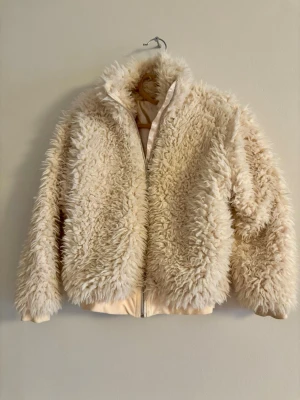 Beige fluffig teddyjacka - Superskön teddyjacka i beige med fluffig, lockig yta och dragkedja framtill. Jackan har hög krage och ribbade muddar nedtill för extra komfort. Perfekt för dig som vill ha en mysig och trendig look i höst och vinter.