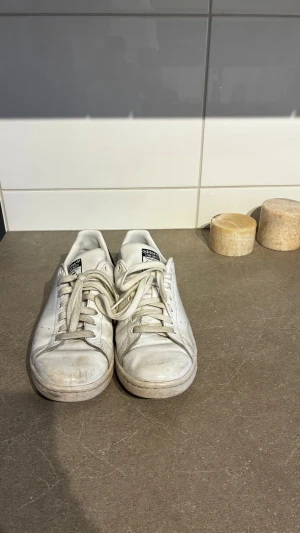 Adidas Stan Smith vita sneakers - Klassiska vita Adidas Stan Smith sneakers med svarta detaljer på hälen och perforerade ränder på sidorna. Skorna har lågt skaft, snörning och är tillverkade i läder med gummisula. Perfekta för en clean och tidlös stil.