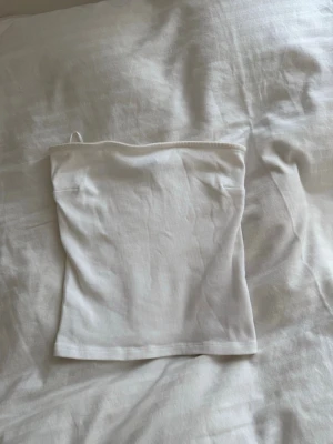 Vit ribbad tubtopp från Gina Tricot - Säljer en enkel och stilren vit ribbad tubtopp från Gina Tricot. Toppen är axelbandslös och har en mjuk, stretchig känsla som passar perfekt till sommaren. Klassisk bandeau-modell som är lätt att matcha med allt.
