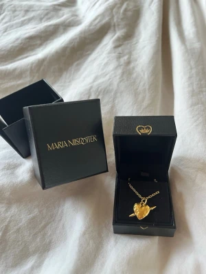 Guldigt hjärthalsband Maria Nilsdotter - Unikt halsband från Maria Nilsdotter i guldtonat material med ett hjärta som hänge. Kedjan är tunn och elegant, och hjärtat har en cool, handgjord känsla. Perfekt för dig som vill ha något personligt och stilrent. I längden 45❤️