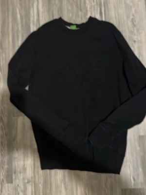 Svart sweatshirt från Hugo Boss - Snygg svart sweatshirt från Hugo Boss med diskret broderad logga på bröstet. Tröjan har rund halsringning och långa ärmar. Tillverkad i mjukt bomullsmaterial som känns skönt mot huden. Perfekt för en clean och stilren look.