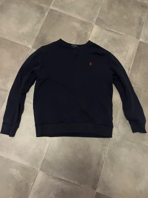 Marinblå sweatshirt från Polo Ralph Lauren - Marinblå sweatshirt från Polo Ralph Lauren med klassisk rund hals och lång ärm. Liten röd broderad logga på bröstet. Perfekt för en avslappnad och stilren look. Mjuk bomullsblandning som känns skön mot huden.