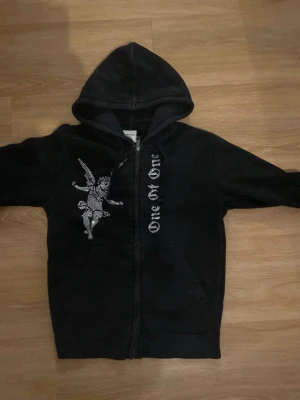 Svart hoodie One of One med ängel - Svart hoodie från One of One med dragkedja, stor huva och fickor framtill. På bröstet finns ett tryck av en ängel och texten 'One of One' i vitt längs dragkedjan. Hoodien har en avslappnad passform och är tillverkad i mjukt material.