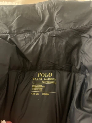 Svart dunjacka från Polo Ralph Lauren - Snygg svart dunjacka från Polo Ralph Lauren Performance med hög krage och dragkedja. Jackan har en puffig design och sidofickor med dragkedja. Perfekt för kalla vinterdagar och riktigt stilren look.