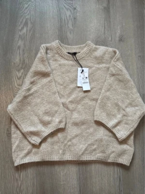 Beige stickad tröja med kort ärm - Beige stickad tröja med korta ärmar och rund halsringning. Tröjan har en avslappnad passform och ribbstickade muddar vid ärmslut och nederkant. Perfekt för lager-på-lager och enkel att matcha med andra plagg.