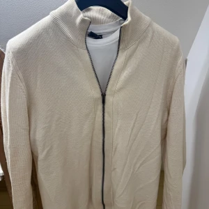Beige ziptröja från Kappahl  - Stilren beige ziptröja från Kappahl med hög krage och diskret strukturstickning. Tröjan har hel dragkedja framtill och ribbade muddar vid halsen. Perfekt att bära över en t-shirt för en clean och avslappnad look.