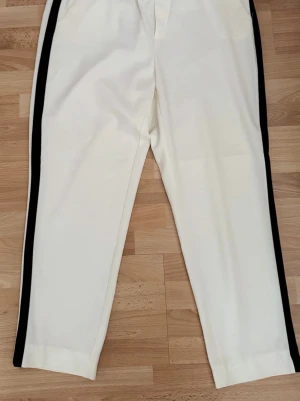Nya. Vita byxor med svarta sidoremsor - Snygga vita byxor med raka ben och svarta sidoremsor längs hela benen. De har en enkel design och är tillverkade i bomull material som ger en bekväm passform. Perfekta för dig som gillar en sportig och stilren look.