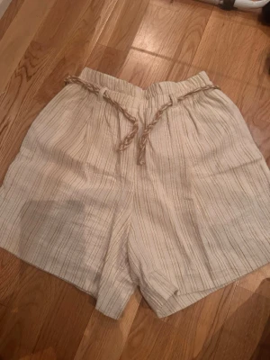 Beige randiga shorts med snörning - Säljer ett par beige shorts med tunna ränder och flätad snörning i midjan. Shortsen har två fickor bak och är tillverkade i ett lätt och luftigt material, perfekta för varma dagar. Passformen är loose och de har en avslappnad vibe.