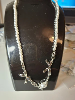 Pärlhalsband med rosetter och strass - Snyggt halsband med vita pärlor, dekorerat med två silvriga rosetter och en stor pärlliknande detalj omgiven av glittrande strass. Kedjan är i silverfärg och ger en cool kontrast till de klassiska pärlorna. Perfekt för dig som gillar att sticka ut med accessoarer.