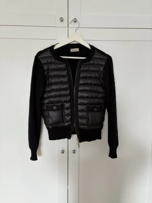 Svart cardiganjacka från Moncler - Snygg svart cardiganjacka från Moncler med quiltad front i glansigt material och stickade ärmar och rygg. Två framfickor med knappstängning och dragkedja. Andvönt sparsamt och har inga defekter. Säljer då den tyvärr it kommer till andvändning längre. Storlek xs men passar mig som har S. Skriv om ni har några frågar❤️